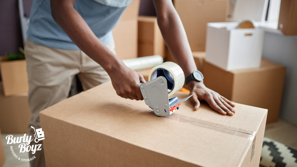 Person taping a moving box in a room full of packed items — packing tips for moving with Burly Boyz Moving & Storage.